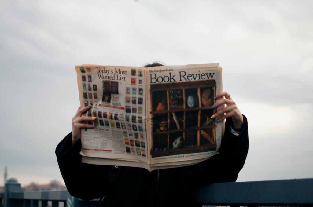 Individual holding a book review newspaper with colorful nails against a cloudy sky backdrop.